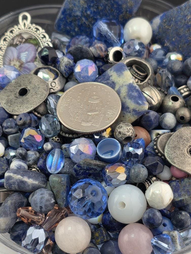 The image is a close-up of a collection of various beads, charms, and coins, all in shades of blue and silver.