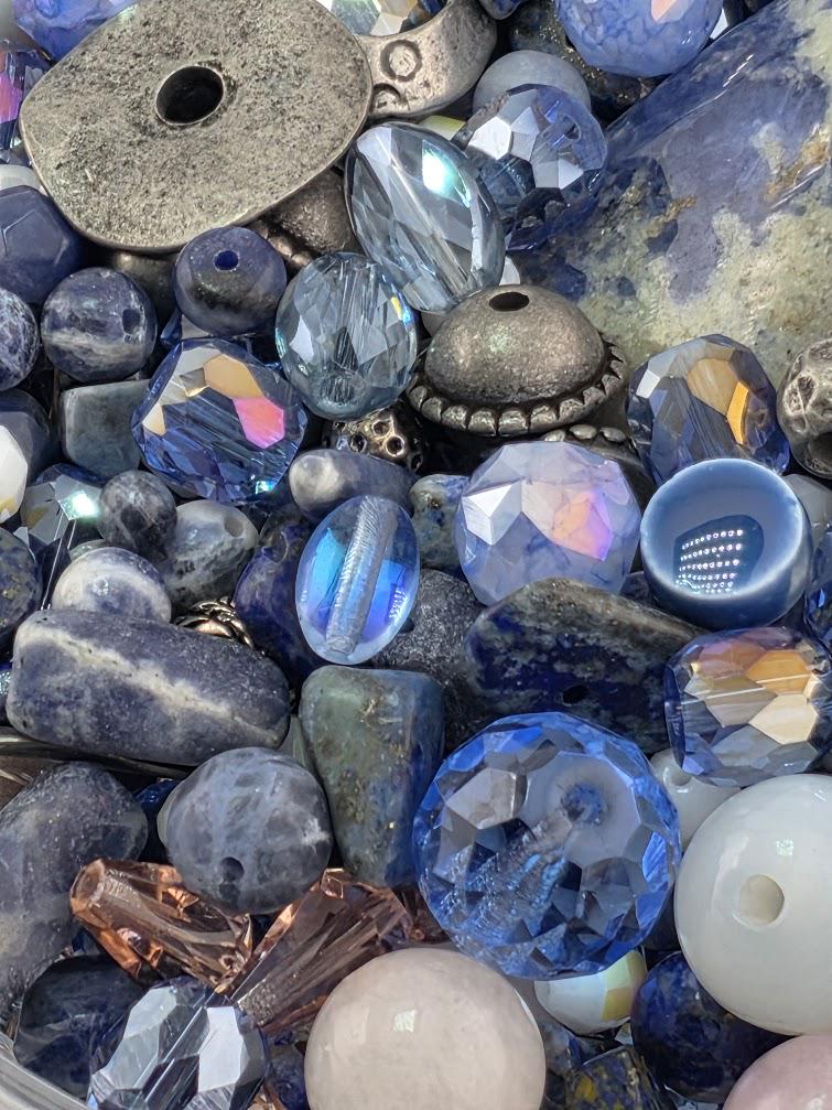 The image is a close-up of various colorful beads and gemstones, including clear, blue, and multicolored stones, as well as metal pieces.