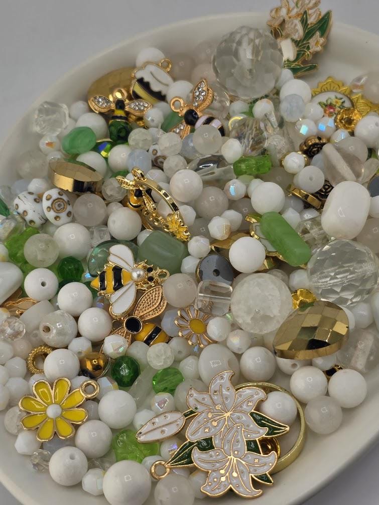 The image is a collection of various beads, charms, and other decorative items, including a bee, flowers, and other embellishments, all arranged on a white surface.
