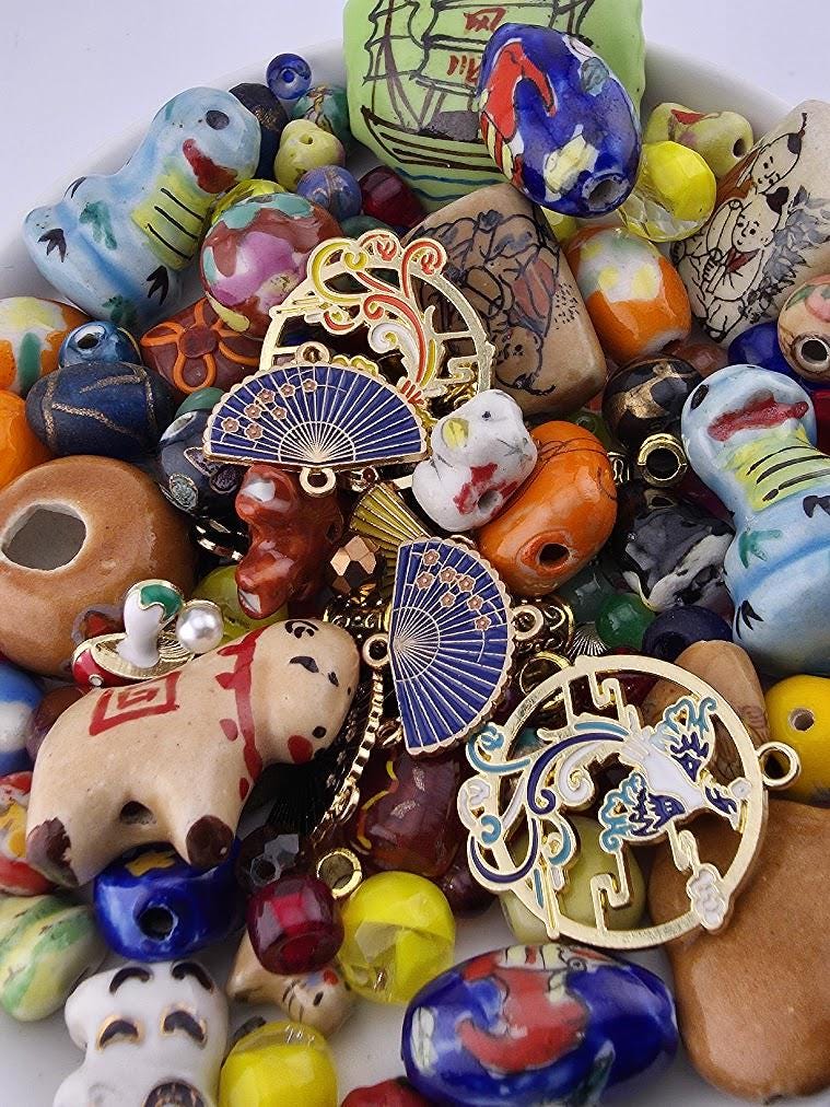 The image is a colorful assortment of various beads, charms, and other decorative items, including a fan-shaped charm, a small elephant charm, and a blue and gold charm.