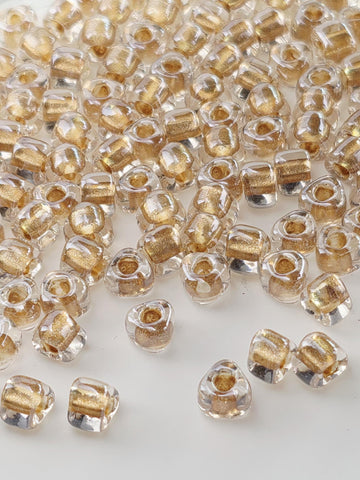 a close-up view of numerous clear glass beads scattered across a white surface. The beads have a golden hue and are arranged in a seemingly random manner, creating a visually striking and intricate pattern.