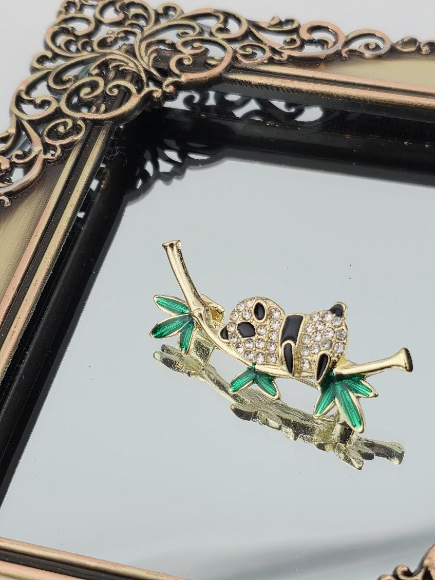 A decorative panda brooch with green leaves and sparkling rhinestones is displayed on a reflective surface, framed by an ornate gold frame.