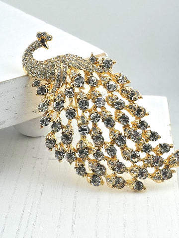 A decorative brooch in the shape of a peacock, adorned with numerous small, clear gemstones in various shades of black and gold.