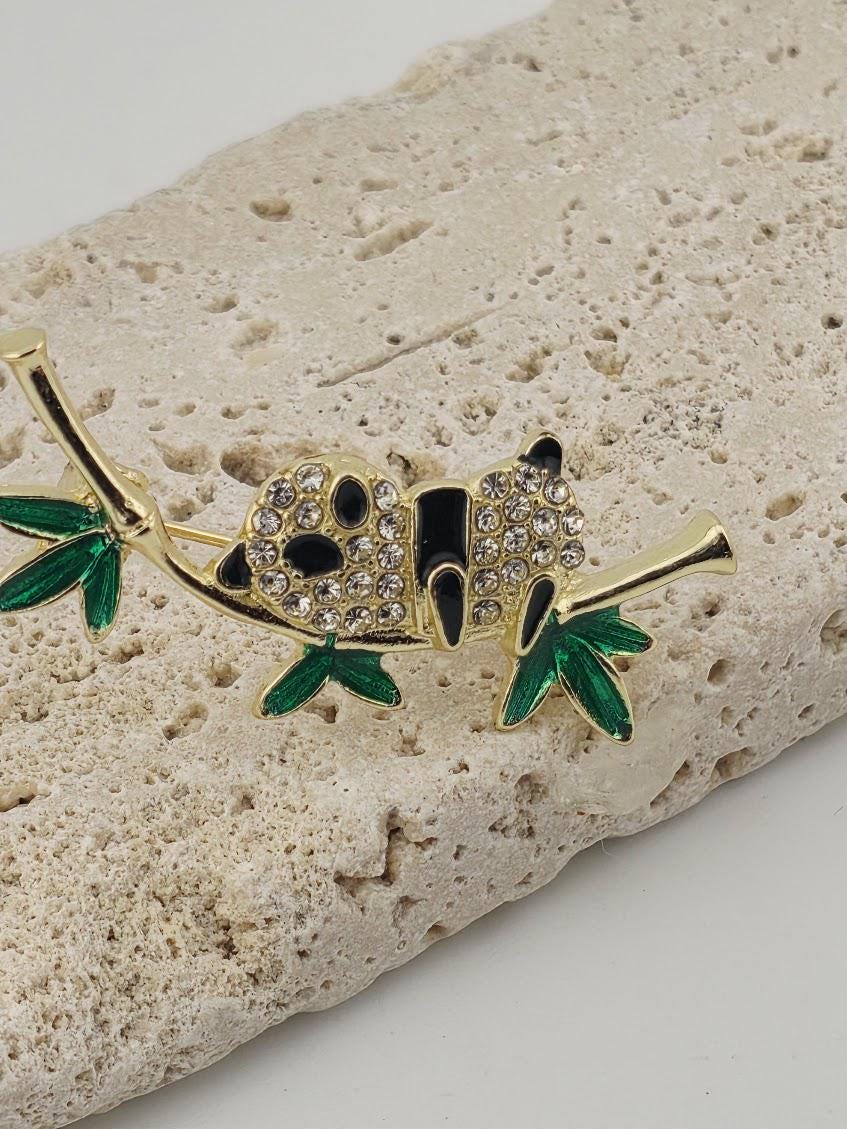 A decorative panda-shaped brooch with green leaves and sparkling rhinestones is displayed on a textured stone surface.