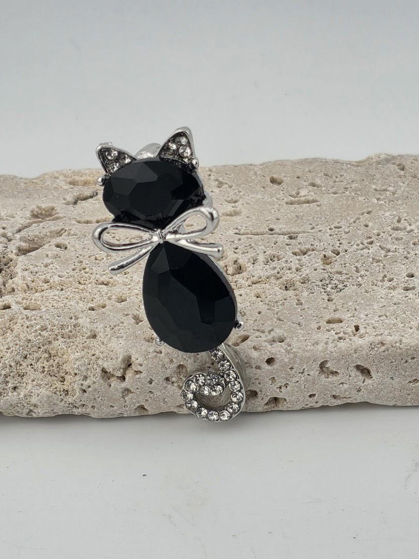 A black cat-shaped pendant with a silver bow and rhinestone accents is displayed on a light-colored stone surface.