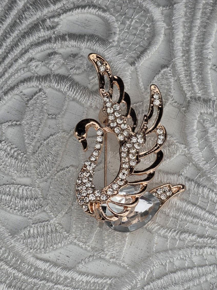 A decorative brooch in the shape of a swan, adorned with sparkling rhinestones, is displayed against a textured gray background.