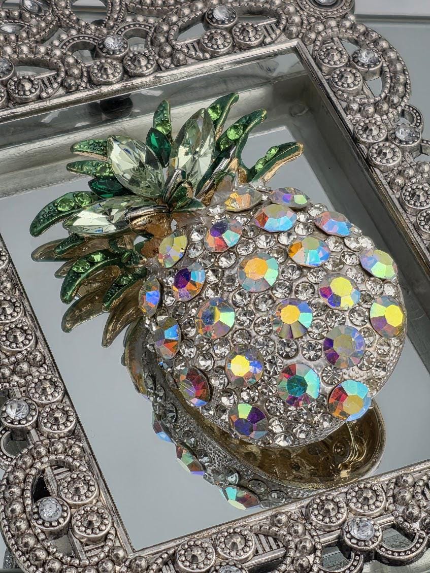a decorative pineapple-shaped piece of jewelry adorned with colorful crystals and gemstones, displayed in a silver frame.