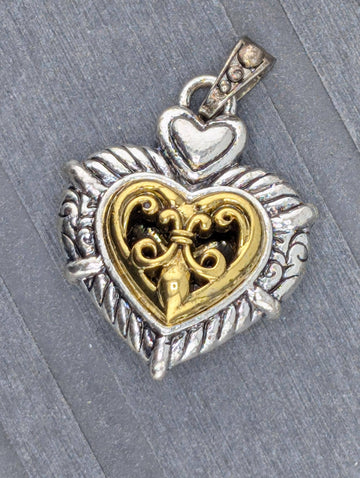 A heart-shaped pendant with a decorative metal design and a small key-like element inside it, resting on a gray surface.