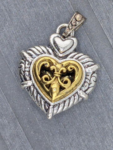 A heart-shaped pendant with a decorative metal design and a small key-like element inside it, resting on a gray surface.