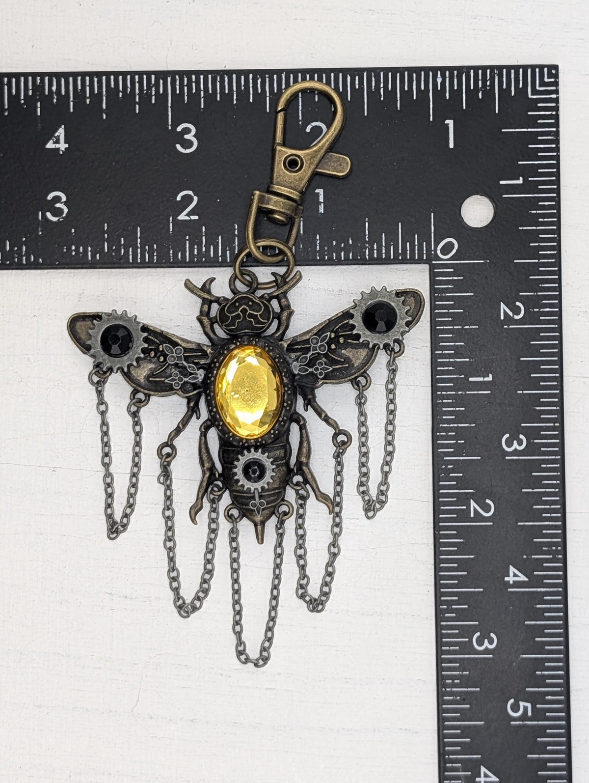A detailed metal keychain in the shape of a bee, with a yellow gemstone in the center and chains hanging from its wings. The keychain is placed next to a ruler for size comparison.