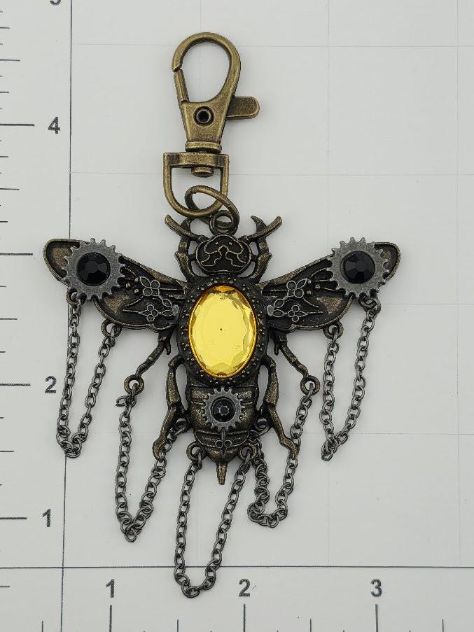 A detailed metal keychain in the shape of a bee, with a yellow gemstone in the center and metal chains hanging from its wings.