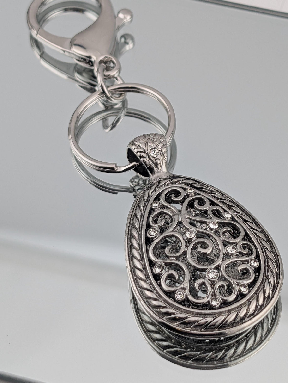 A silver-colored keychain with an ornate, decorative pendant featuring a swirling pattern and small, clear stones.
