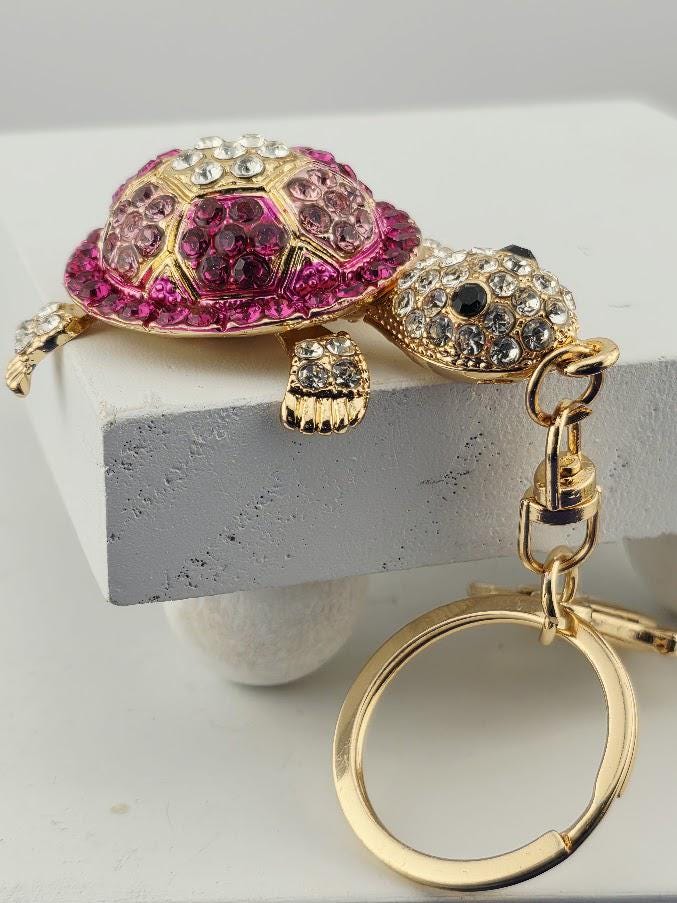 A keychain with a pink and gold turtle design, adorned with rhinestones and a gold ring.