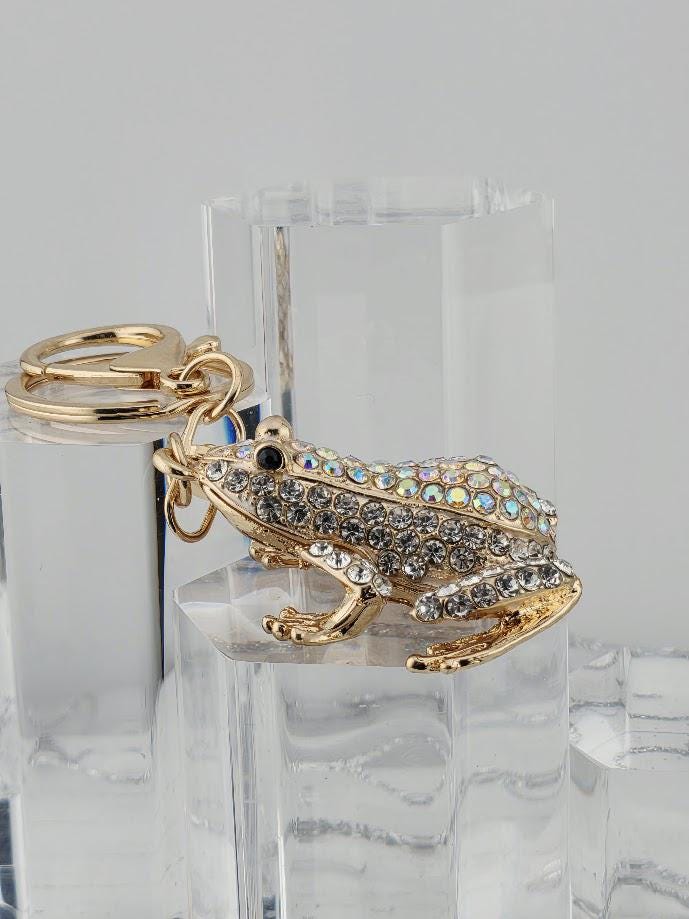 A gold-colored keychain in the shape of a frog, adorned with rhinestones or crystals, is displayed on a clear glass stand.
