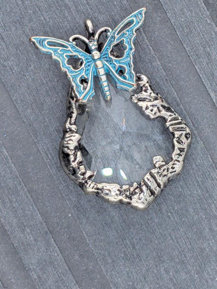A silver butterfly pendant with a clear gemstone in the center, set against a gray background.