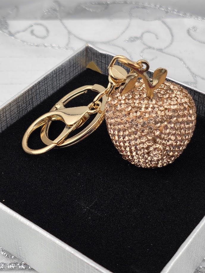 A gold-colored keychain in the shape of a small apple, adorned with rhinestones or crystals, is the focal point of the image.
