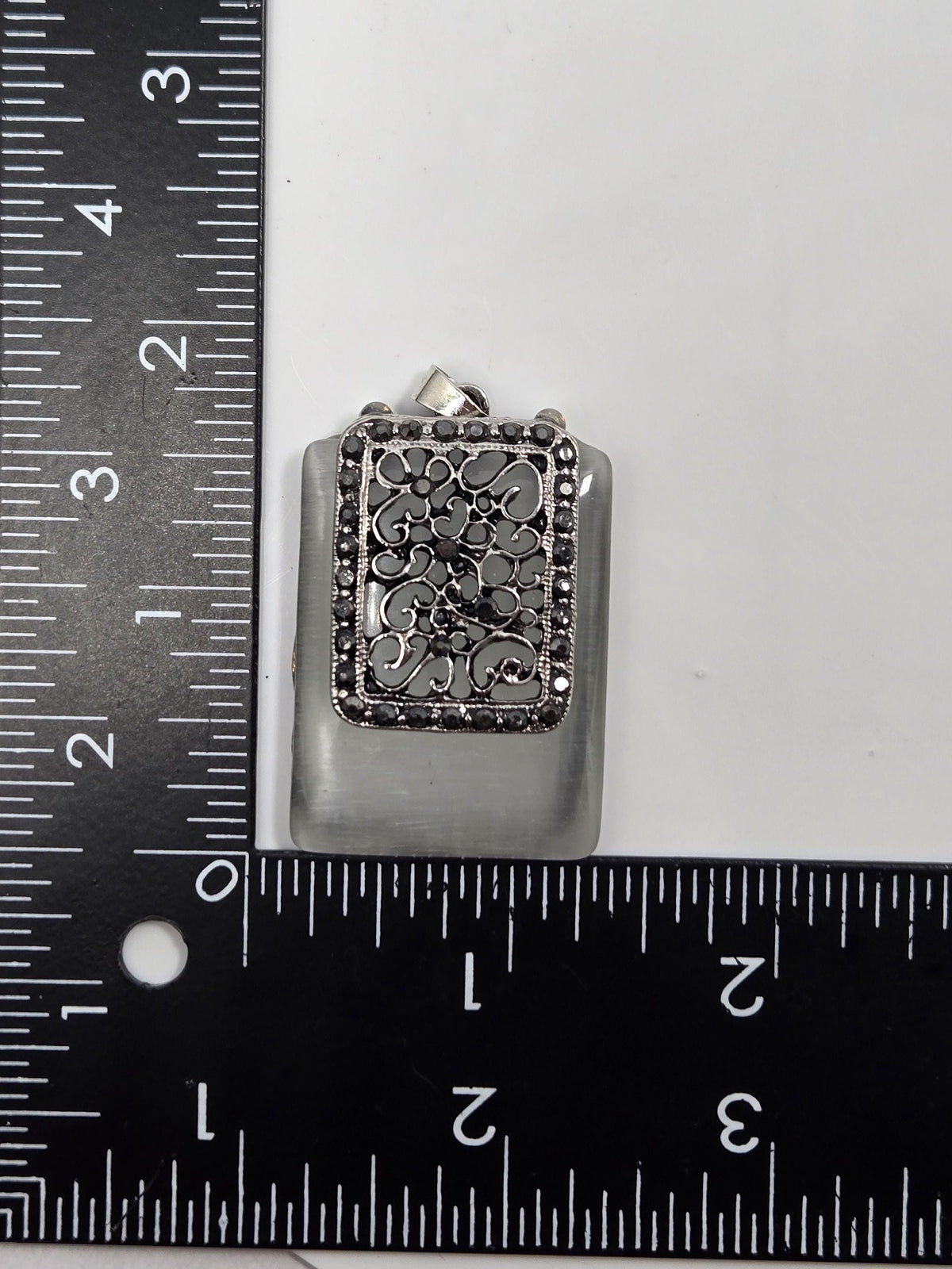 a small, ornate metal clasp or clasp attached to a piece of jewelry, placed next to a ruler for size comparison.