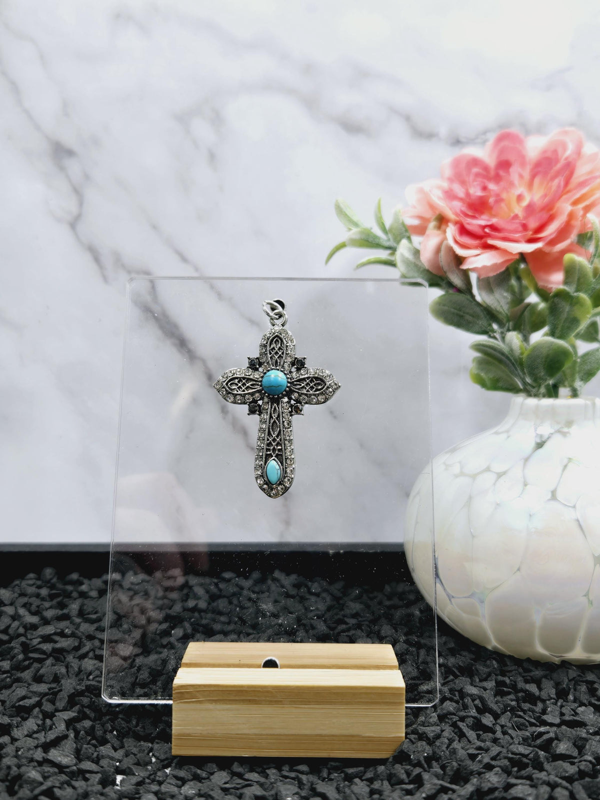 a silver cross pendant with turquoise stones, placed on a marble surface next to a vase with pink flowers and a wooden stand.