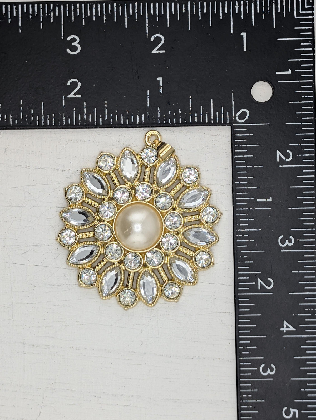 a gold-colored pendant with a pearl at its center, surrounded by numerous clear gemstones. The pendant is placed next to a ruler for size comparison.