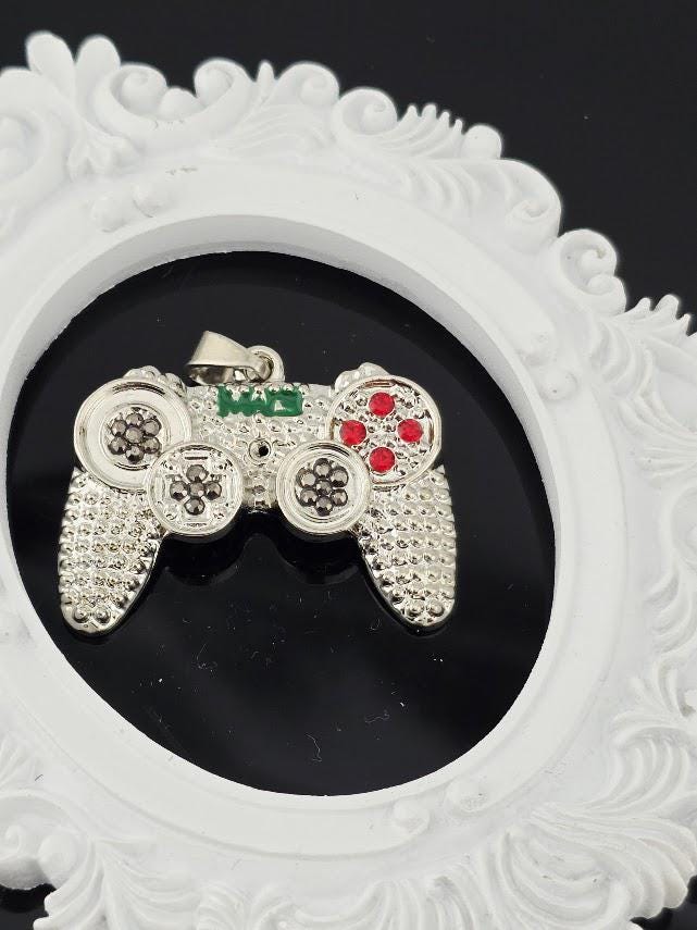 A silver pendant in the shape of a video game controller, with red buttons and a green joystick, is displayed against a white ornate frame.