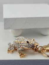 A gold-colored frog keychain with rhinestones or crystals on it, placed on a white surface.