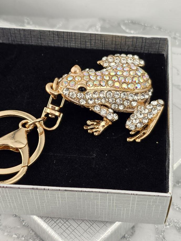 A gold-colored frog keychain with rhinestones or crystals on it, placed on a white surface.