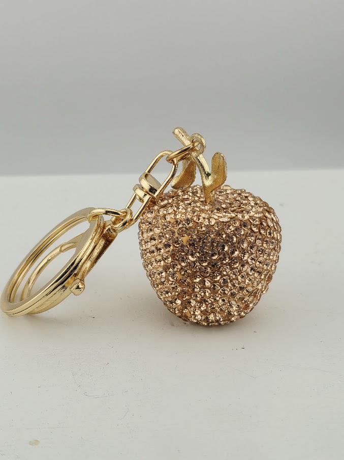 A gold-colored keychain in the shape of a small apple, adorned with rhinestones or crystals, is the focal point of the image.