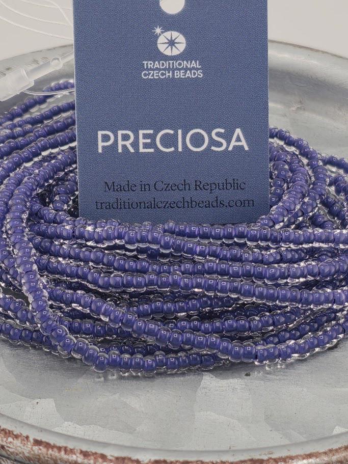 a close-up view of a pile of purple beads on a surface, with a tag attached to it that reads "PRECIOSA" and "Made in Czech Republic".