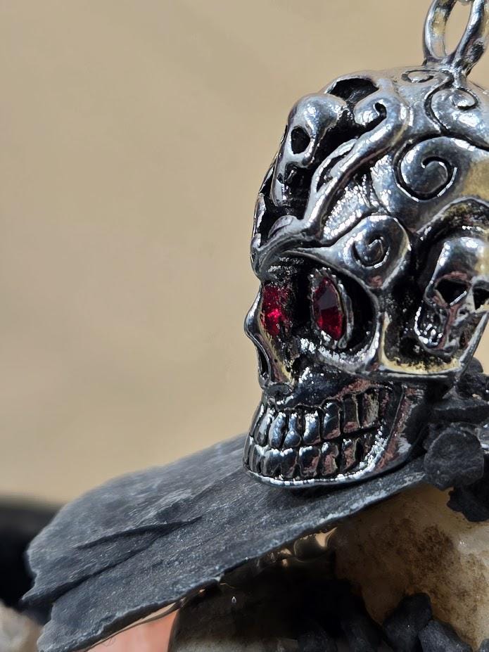 A close-up view of a silver skull pendant with red gemstones, set against a beige background.