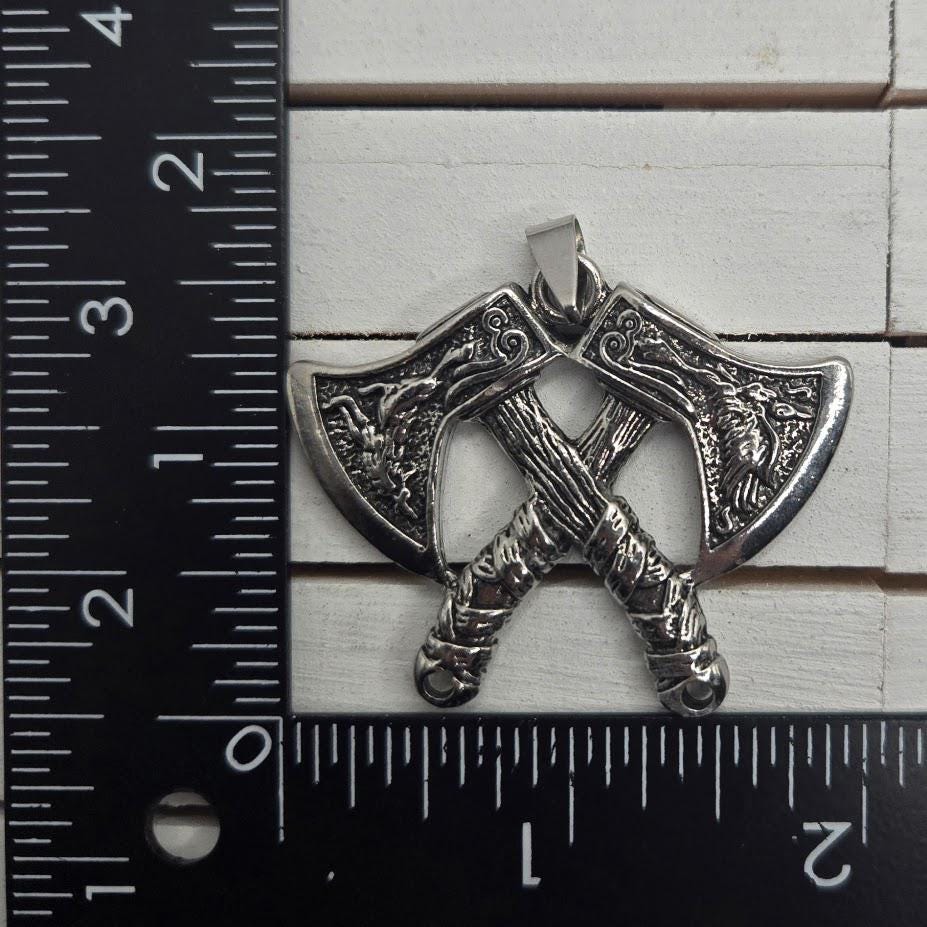 A silver-colored pendant in the shape of a crossed sword or axe, with intricate designs and measurements on a ruler.