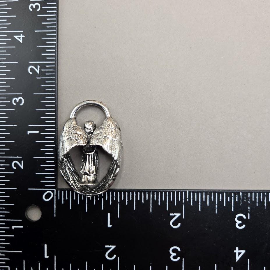 A silver-colored metal charm or pendant with wings is placed next to a ruler on a gray surface.