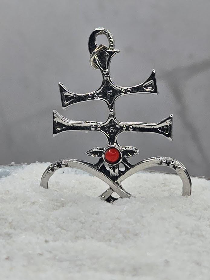 A silver pendant with a red gemstone, featuring an intricate design, is displayed on a bed of white sand.