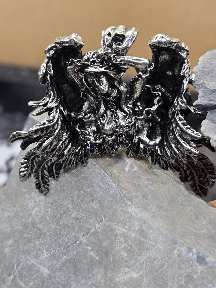 A close-up image of a silver, sculpted pendant or brooch with intricate, winged design elements.
