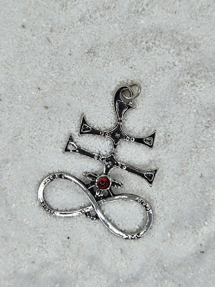 A silver pendant with a cross and a red gemstone, lying on a sandy surface.