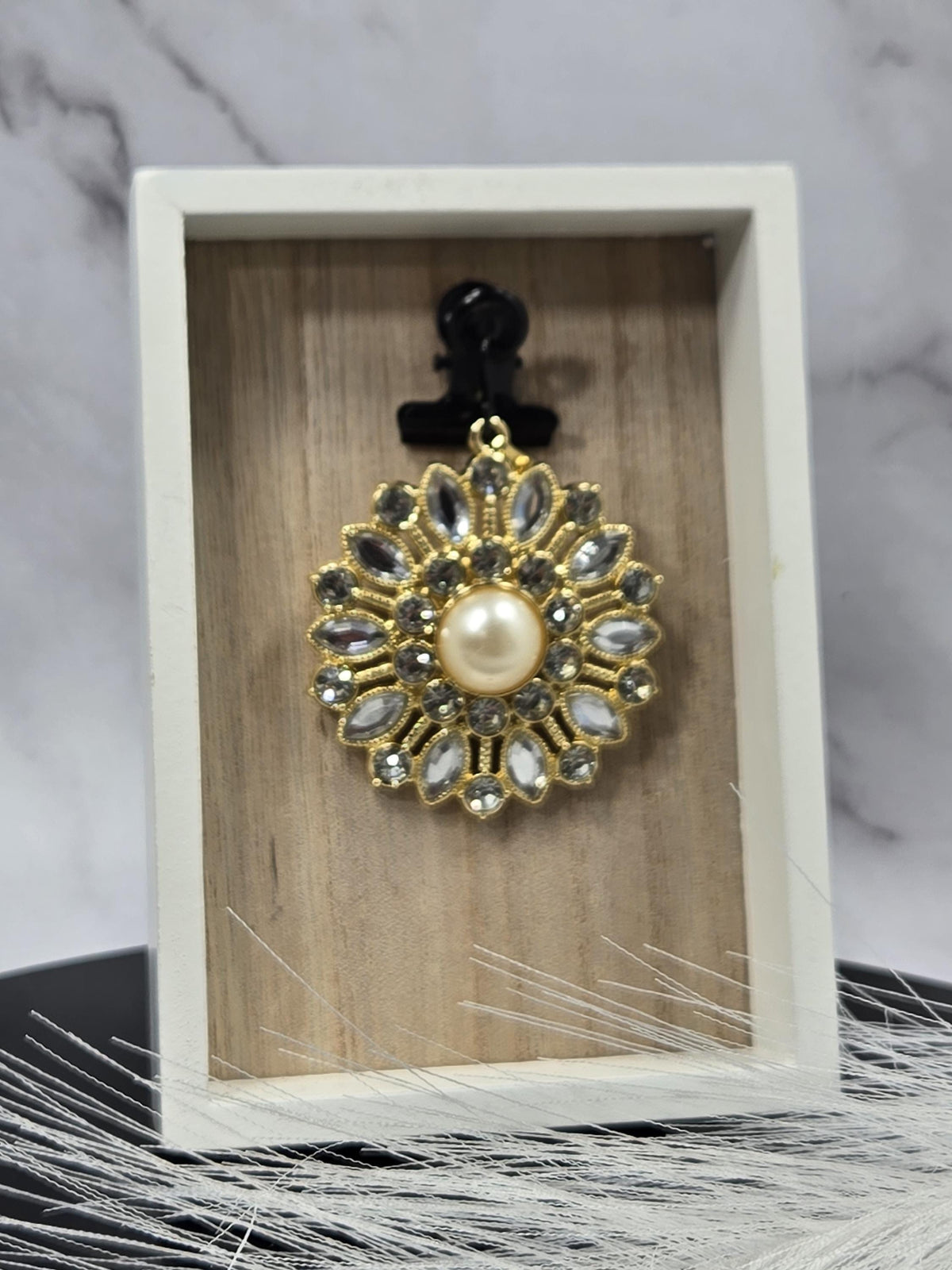 A decorative brooch with a pearl at its center, surrounded by a floral design, is displayed in a white box.