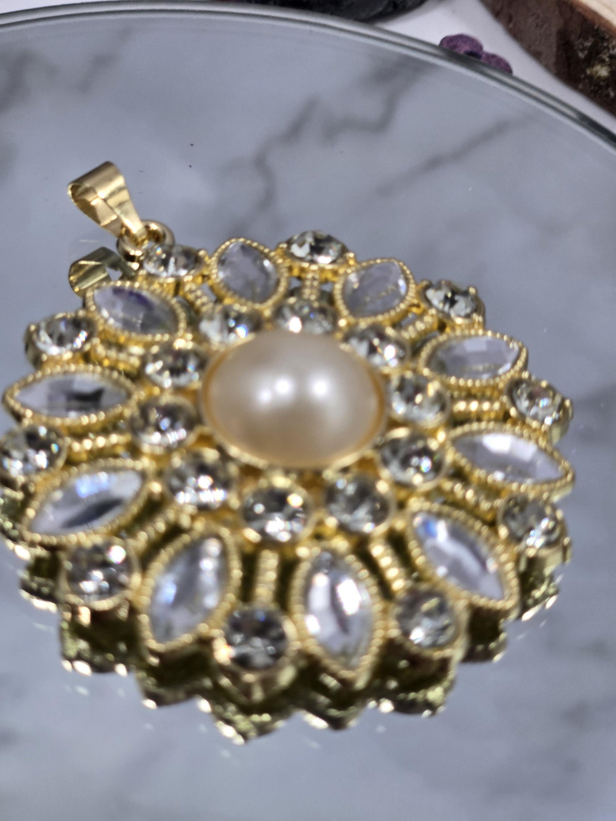 A close-up view of a decorative pendant or brooch with a large pearl at its center, surrounded by numerous small, clear gemstones or crystals in a gold-colored metal frame.
