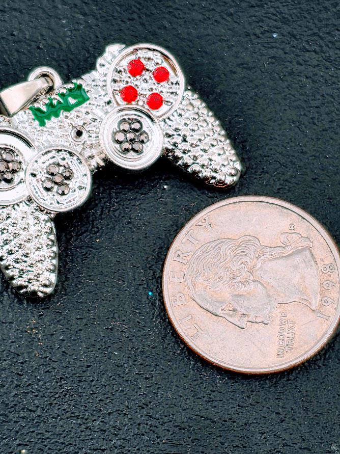 A silver-colored pendant in the shape of a video game controller, adorned with red and green gemstones, is placed next to a United States quarter on a dark surface.