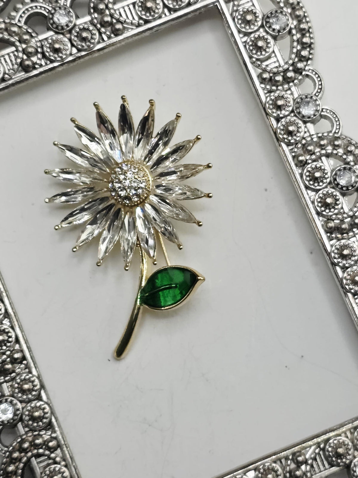 A decorative brooch with a large crystal flower and a green leaf is displayed against a white background, framed by an ornate silver frame.