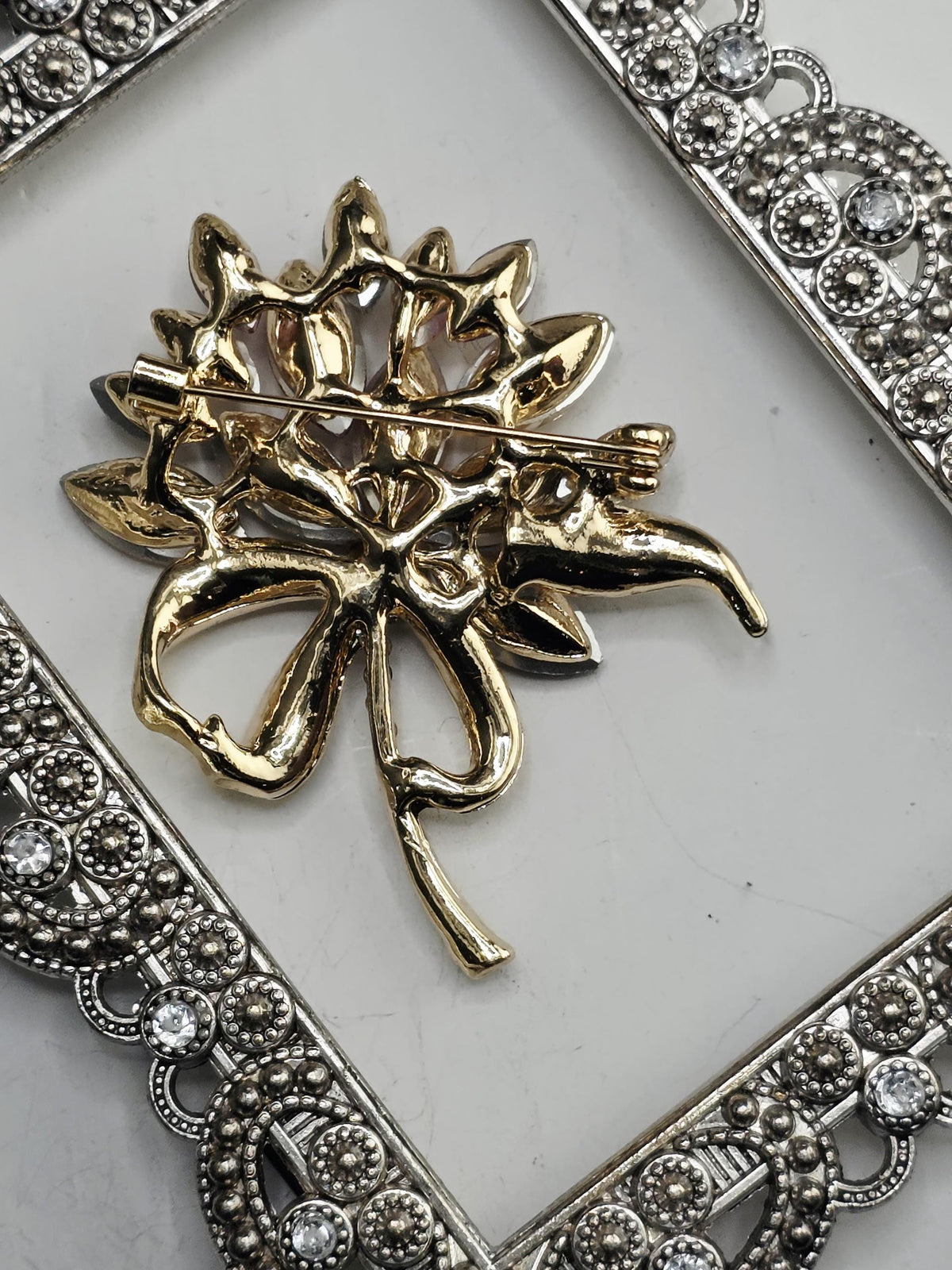 A decorative gold brooch with a floral design is displayed against a white background, framed by an ornate silver frame.