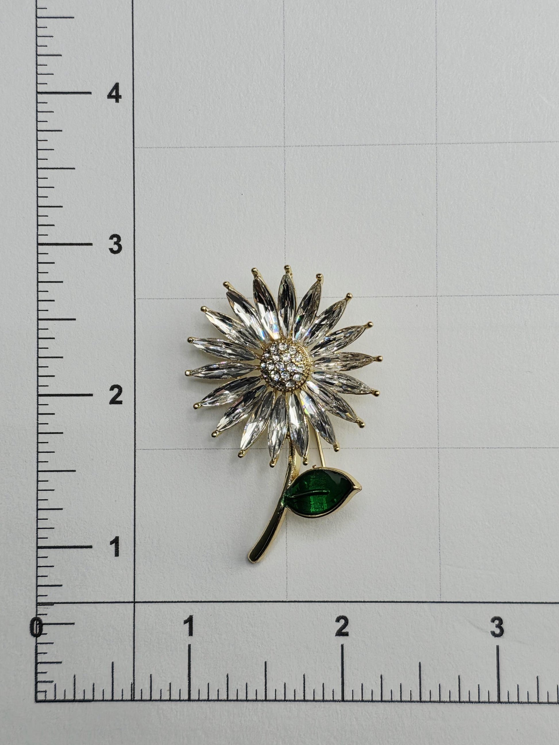 a decorative brooch with a flower-like design and a green leaf-like element, placed on a white surface with a ruler for scale.