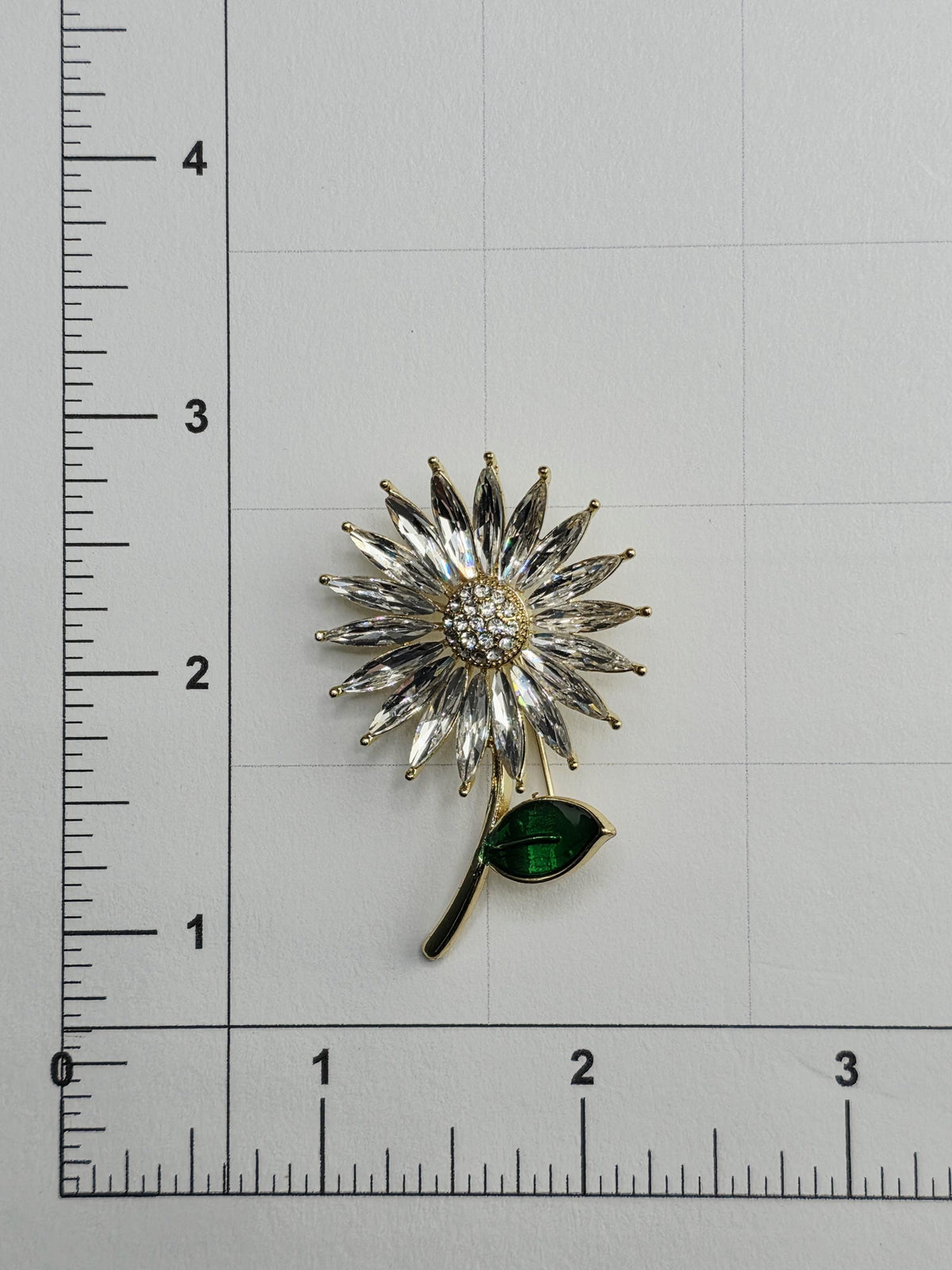 a decorative brooch with a flower-like design and a green leaf-like element, placed on a white surface with a ruler for scale.