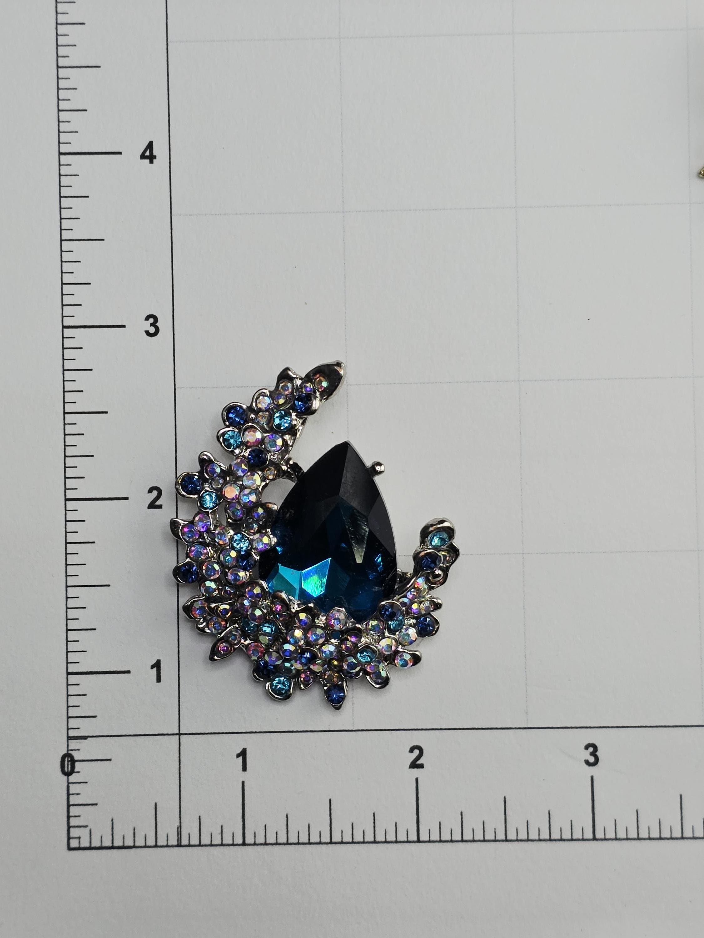 a close-up view of a decorative piece, possibly a brooch or pendant, with a large blue gemstone surrounded by smaller blue and purple gemstones. The piece is placed on a ruler for scale, indicating its size.