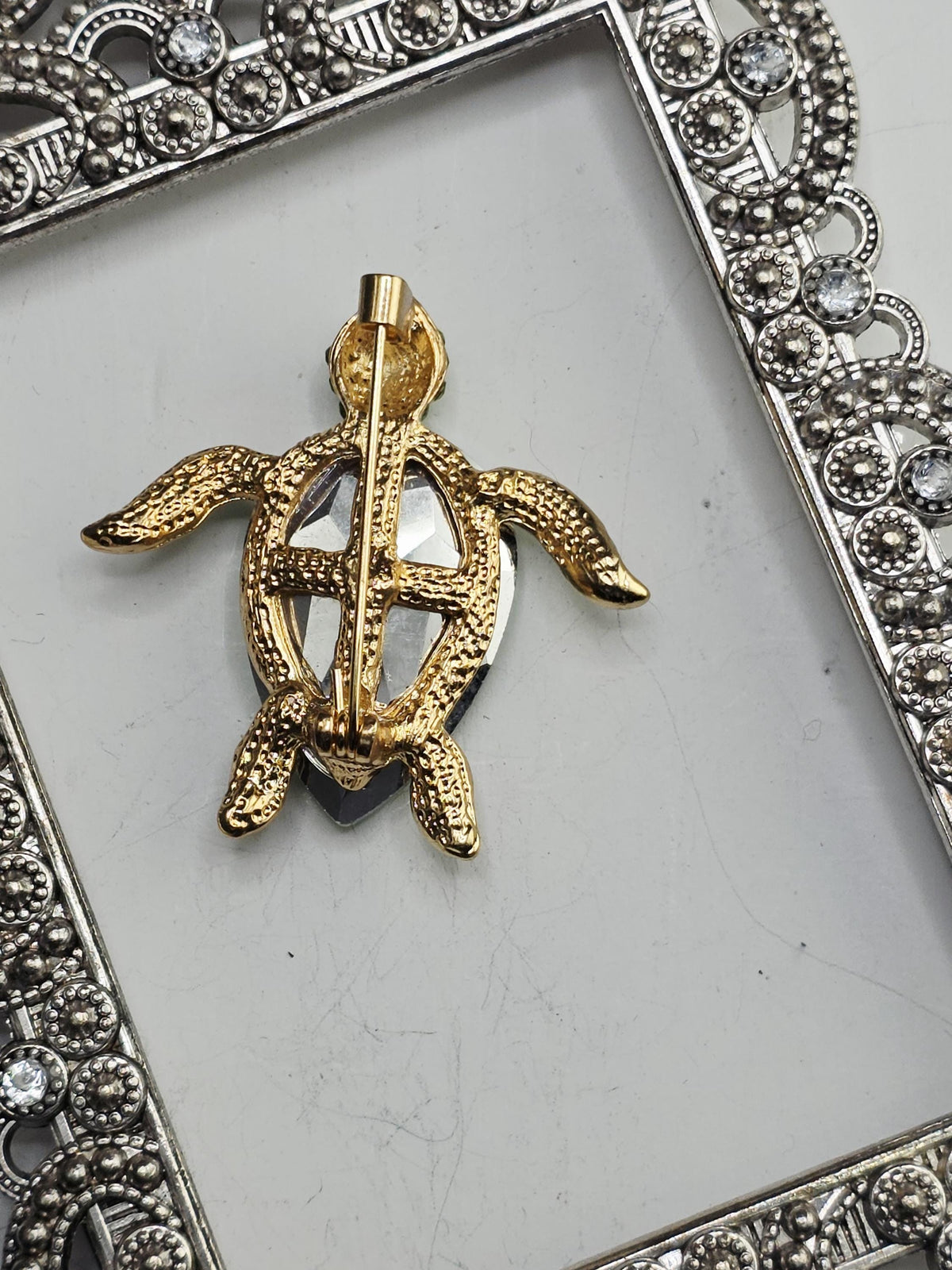 A gold-colored pin or brooch in the shape of a turtle, displayed on a white surface within an ornate metal frame.