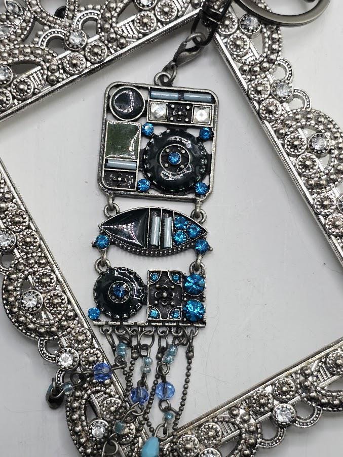a decorative pendant with a combination of blue and silver elements, including a square-shaped design with various shapes and patterns, as well as a hanging chain with blue and silver beads. The pendant is set within an ornate metal frame, which adds to its overall aesthetic appeal.