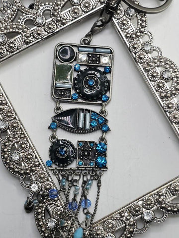a decorative pendant with a combination of blue and silver elements, including a square-shaped design with various shapes and patterns, as well as a hanging chain with blue and silver beads. The pendant is set within an ornate metal frame, which adds to its overall aesthetic appeal.