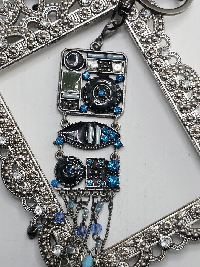 a decorative pendant with a combination of blue and silver elements, including a square-shaped design with various shapes and patterns, as well as a hanging chain with blue and silver beads. The pendant is set within an ornate metal frame, which adds to its overall aesthetic appeal.