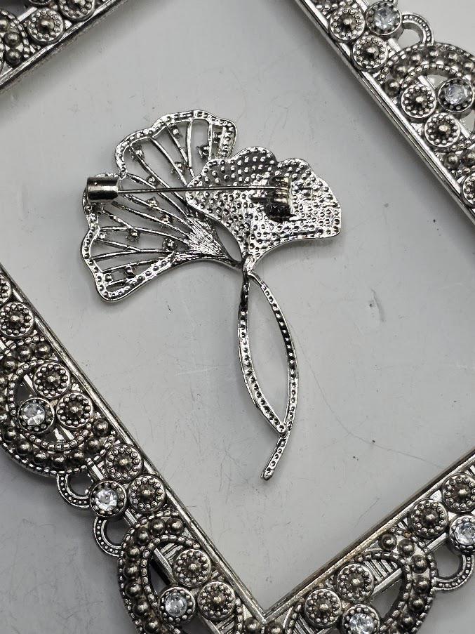 a decorative silver brooch with a floral design, placed on a white surface within an ornate frame.