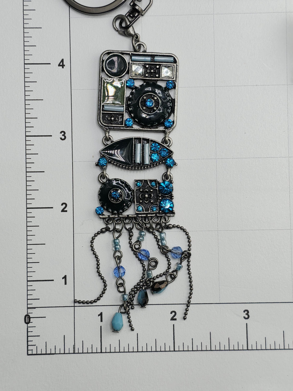A decorative metal bracelet with various charms and beads, including blue and black elements, is shown on a ruler for size comparison.