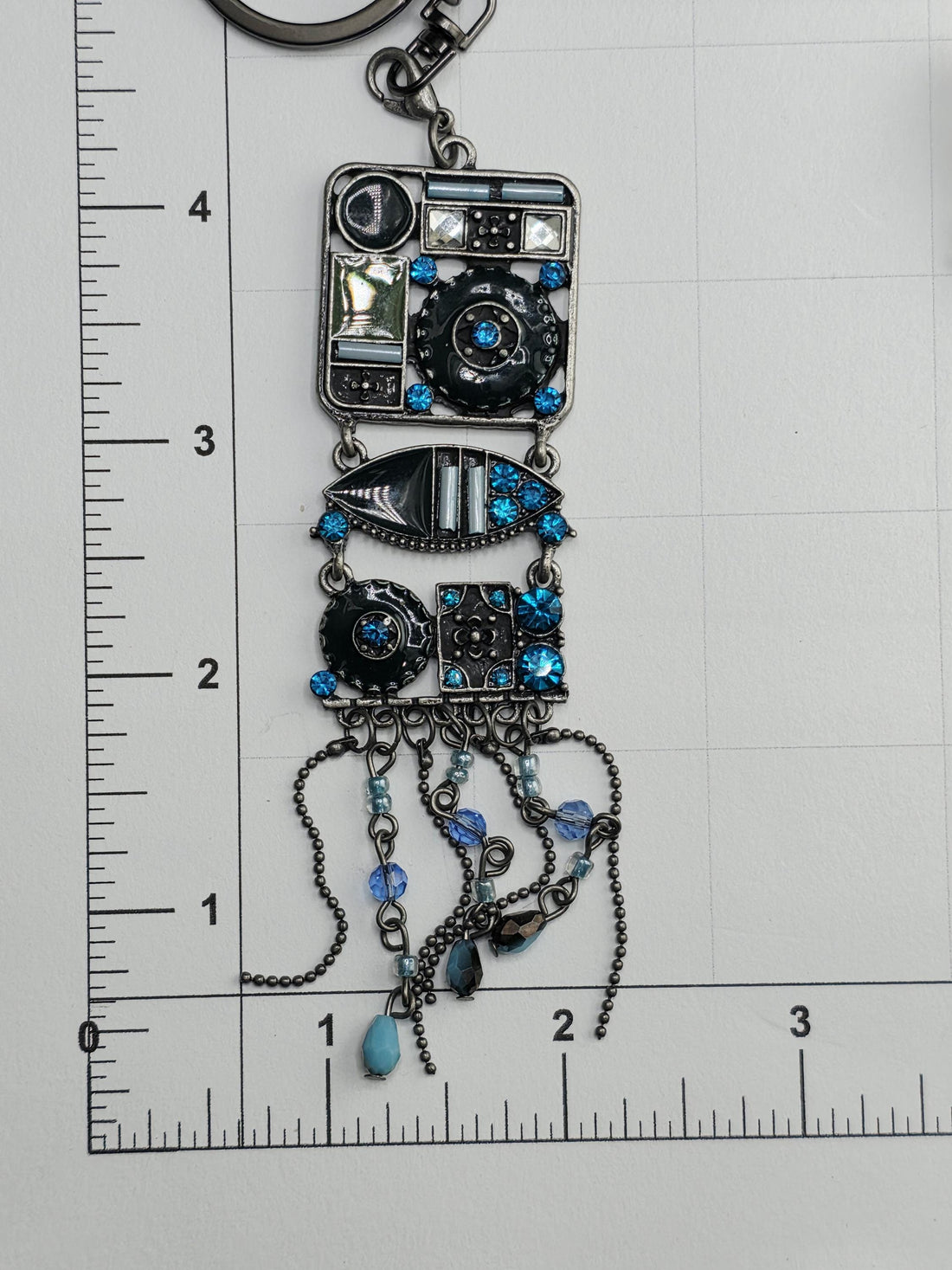 a decorative pendant with a combination of blue and silver elements, including a square-shaped design with various shapes and patterns, as well as a hanging chain with blue and silver beads. The pendant is set within an ornate metal frame, which adds to its overall aesthetic appeal.