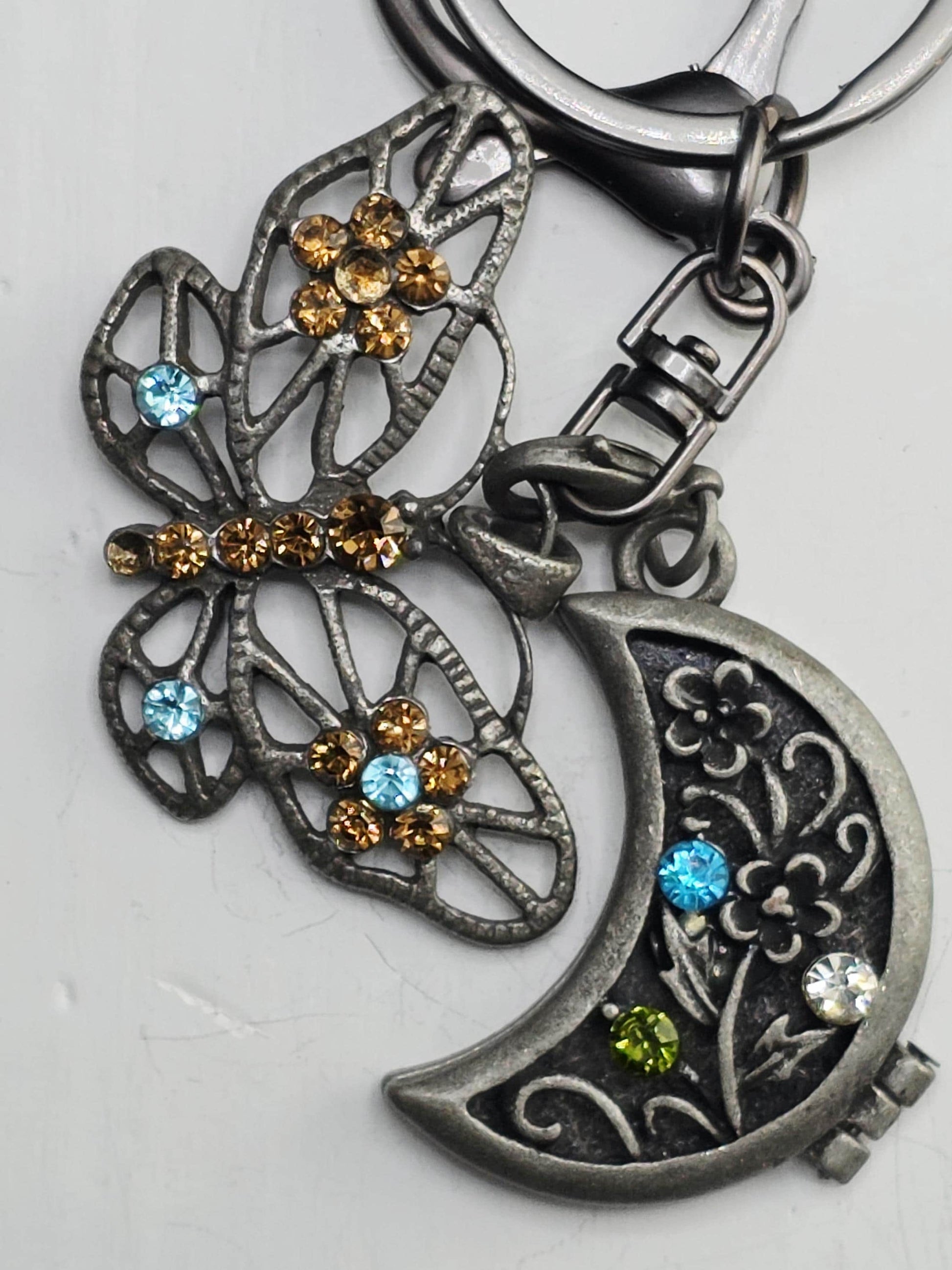 A decorative keychain with a butterfly design and a crescent moon charm. The butterfly has a metallic frame with colorful gemstones, and the crescent moon charm is adorned with blue and green gemstones.